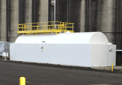 Large white storage tank with a yellow safety railing, situated on a paved surface near industrial structures