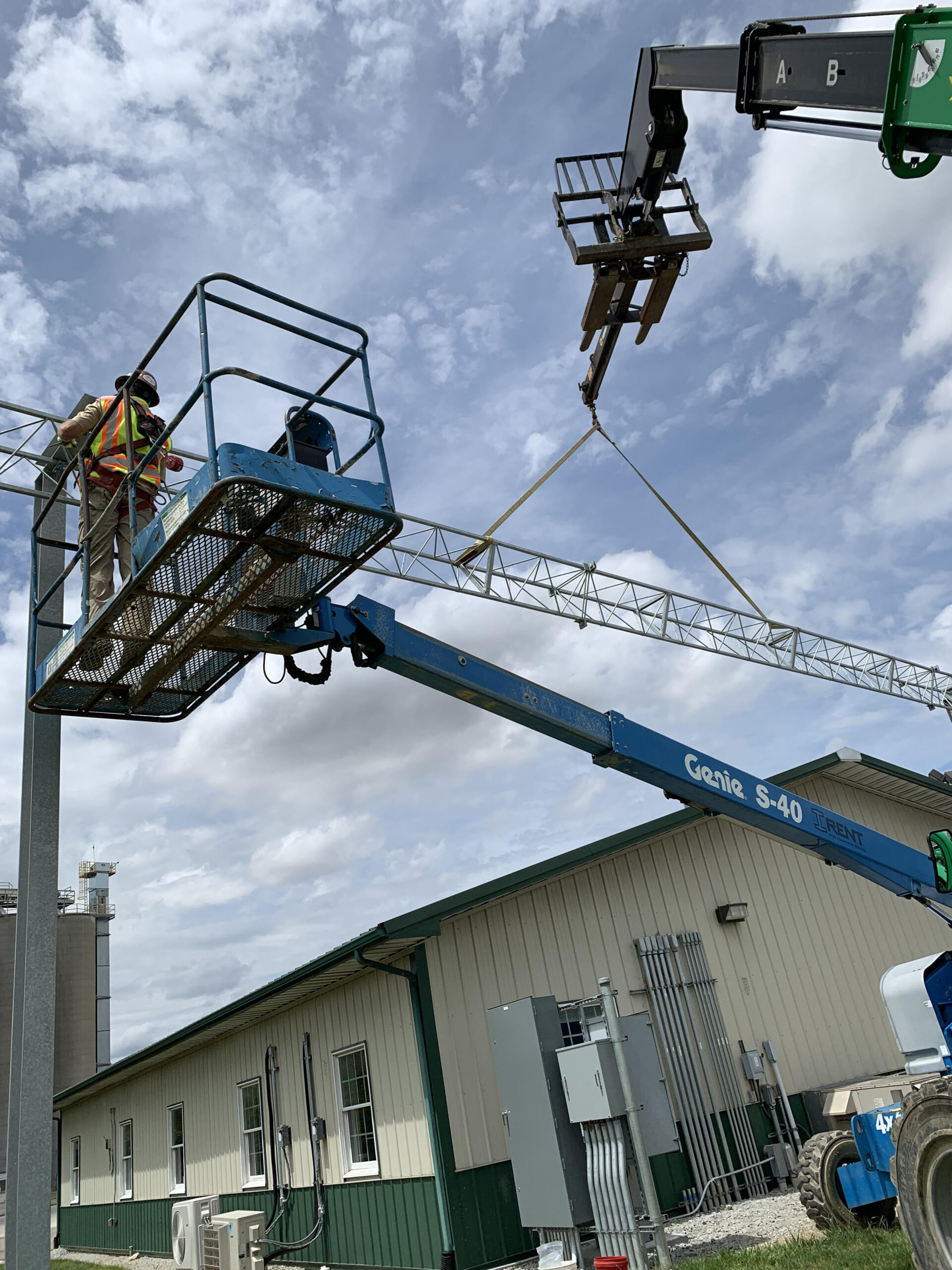 A worker in a safety vest operates a Genie S-40 lift to position a metal truss in a construction setting under a cloudy sky
